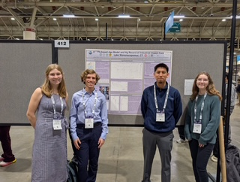 Four individuals stand in front of a scientific poster titled “Utilizing the Global Learning Observations to Benefit the Environment (GLOBE) Tool for Lake Microplastic Research.” They wear conference badges and pose in a convention center surrounded by other posters and attendees. The poster includes text, graphs, and tables, highlighting student-led research on microplastic pollution in lakes using the GLOBE tool.