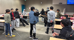 A group of individuals stands in a classroom, participating in a hands-on activity involving straw-based cantilever construction. Several people are arranged in a semi-circle, connecting straws in a web-like structure. One person is in the center, interacting with the structure while others observe or assist. Tables and chairs surround the group, and some participants are seated. The scene reflects a collaborative learning exercise focused on engineering, problem-solving, and communication.
